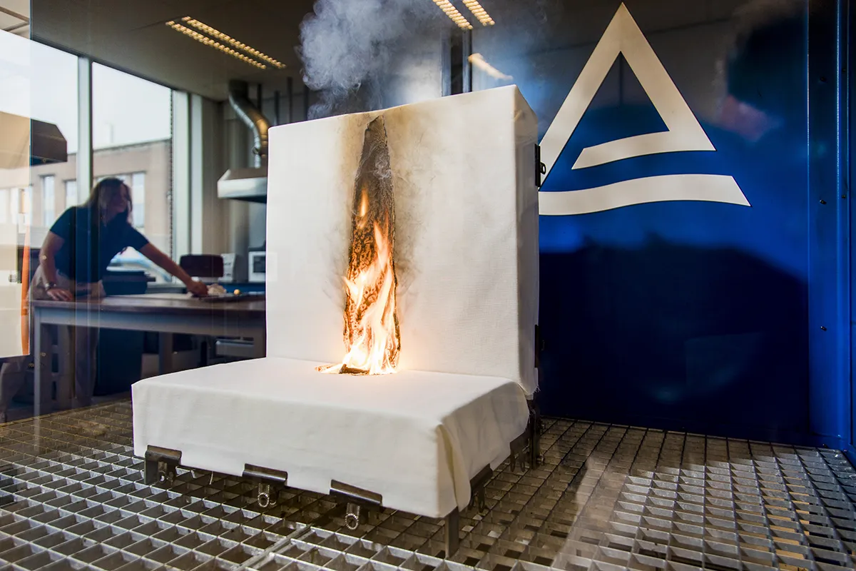 Een gecontroleerde brandtest vindt plaats in een laboratorium waarbij in het midden een wit materiaal brandt, waardoor rook vrijkomt. De opstelling bevindt zich op een metalen roosteroppervlak onder een brandveiligheidskap. Op de achtergrond lijkt het alsof een persoon apparatuur bedient of aantekeningen maakt in een laboratorium met glazen wanden.