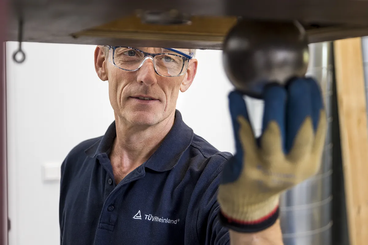 Een man met een veiligheidsbril, een blauw poloshirt met bedrijfslogo en beschermende handschoenen houdt een metalen voorwerp vast in een laboratorium of industriële omgeving. Hij kijkt aandachtig naar het object, dat deel uitmaakt van een mechanisch of testapparaat boven hem.