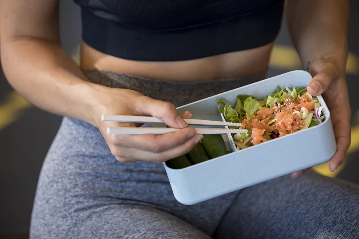 Een persoon in trainingskleding zit en gebruikt eetstokjes om een gezonde maaltijd uit een rechthoekige bak te eten. De maaltijd bestaat uit zalm, gemengde groenten en komkommers. De persoon draagt een zwart sportshirt en een grijze legging. De achtergrond is wazig, waardoor de nadruk op de persoon en de maaltijd ligt: een scène die perfect is voor bedrijfsfotografie.