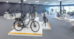 Een moderne showroom ontworpen voor bedrijfsfotografie toont meerdere fietsen, met prominente e-bikes van het merk Amslod op verhoogde witte platforms met gele bies. De kamer heeft grijze muren, grote ramen aan één kant die natuurlijk licht binnenlaten, en een zithoek met witte stoelen en een ronde tafel in de hoek.