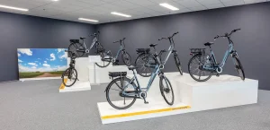 De afbeelding toont een showroom met zes moderne fietsen op witte presentatieplatforms tegen een grijze muur. Aan de ene kant versterkt een grote gedrukte foto van een schilderachtige weg en een blauwe lucht het tafereel. De fietsen, opgesteld op verschillende hoogtes, hebben een strak design met geïntegreerde verlichting en bagagedragers, ideaal voor bedrijfsfotografie.