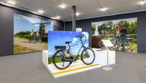 Foto van een showroom met elektrische fietsen. Een zilveren elektrische fiets wordt prominent tentoongesteld op een witte standaard met op de achtergrond een landelijk tafereel. Grote posters erachter tonen schilderachtige fietsroutes, waaronder een met een windmolen en een andere met twee fietsende mensen bij een fontein – perfect voor bedrijfsfotografie.