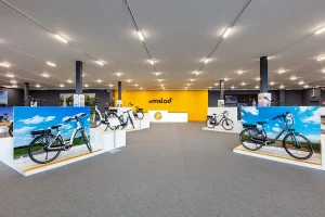 Een moderne showroom met een felgele muur met centraal het 'amslod'-logo. De ruimte, perfect voor bedrijfsfotografie, beschikt over meerdere elektrische fietsen op witte platforms met daarachter grote schilderachtige foto's van luchten en velden. Plafondlampen creëren een goed verlicht interieur met vloerbedekking en extra zichtbare tentoonstellingsruimtes.