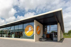 Een modern commercieel gebouw met grote ramen met twee ronde logo's, waarop een wit paard in beweging is afgebeeld op een gele achtergrond. Het bouwwerk heeft een overhangend dak, een verharde entree en meerdere fietsen geparkeerd bij de ingang. De lucht is gedeeltelijk bewolkt, perfect voor bedrijfsfotografie.