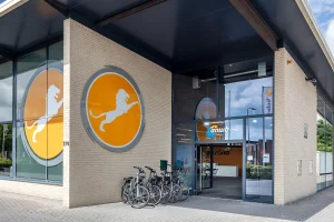 Een modern gebouw met beige bakstenen muren heeft aan de buitenkant grote ronde logo's van een gele leeuw. De entree heeft glazen deuren en een bord met de tekst ‘ANWB’. Er staan meerdere fietsen buiten geparkeerd, waardoor ideale bedrijfsfotografie mogelijk is. Het gebied is goed verlicht en de lucht is gedeeltelijk bewolkt.