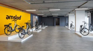 Een strakke showroom heeft aan de linkerkant een felgele muur waarop het zwarte 'amslod'-logo prominent zichtbaar is. Op witte platforms rondom de ruimte staan diverse fietsen netjes tentoongesteld. De grijze vloer en zichtbare buizen aan het plafond dragen bij aan het moderne, industriële ontwerp, perfect voor bedrijfsfotografie.