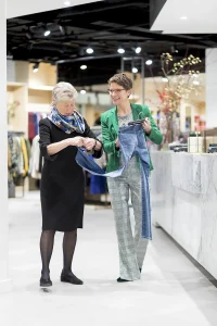 Twee vrouwen staan in een lichte, moderne kledingwinkel. Een vrouw, met kort grijs haar en een kleurrijke sjaal, onderzoekt een spijkerbroek. De andere vrouw, met een bril en een groene blazer over een wit overhemd met een geruite broek, houdt de spijkerbroek omhoog en glimlacht. Op de achtergrond zijn kledingrekken zichtbaar: een perfect decor voor bedrijfsfotografie.