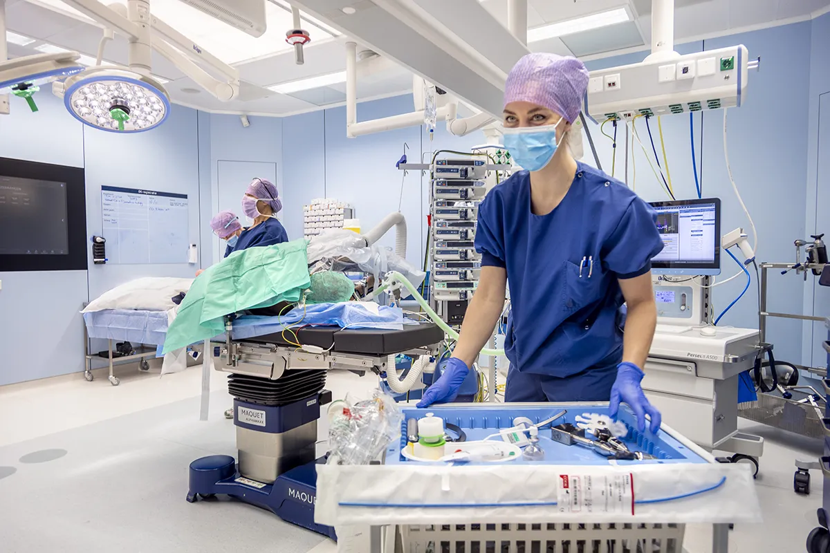 Een medische professional met blauwe scrubs, een chirurgisch masker en een paarse pet staat in een operatiekamer naast een dienblad met chirurgische instrumenten. Een ander personeelslid op de achtergrond is bezig met het afstellen van de apparatuur bij de operatietafel. De ruimte, vastgelegd via bedrijfsfotografie, is goed verlicht en er zijn monitoren en medische apparatuur aanwezig.