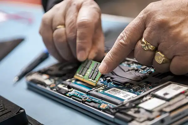 Close-up van handen, versierd met ringen, die voorzichtig een groene RAM-chip in het moederbord van een laptop installeren. De gedeeltelijk gedemonteerde laptop onthult verschillende interne componenten en circuits. Dit nauwgezette proces toont de precisie van een bedrijfsfotograaf die elk ingewikkeld detail vastlegt.