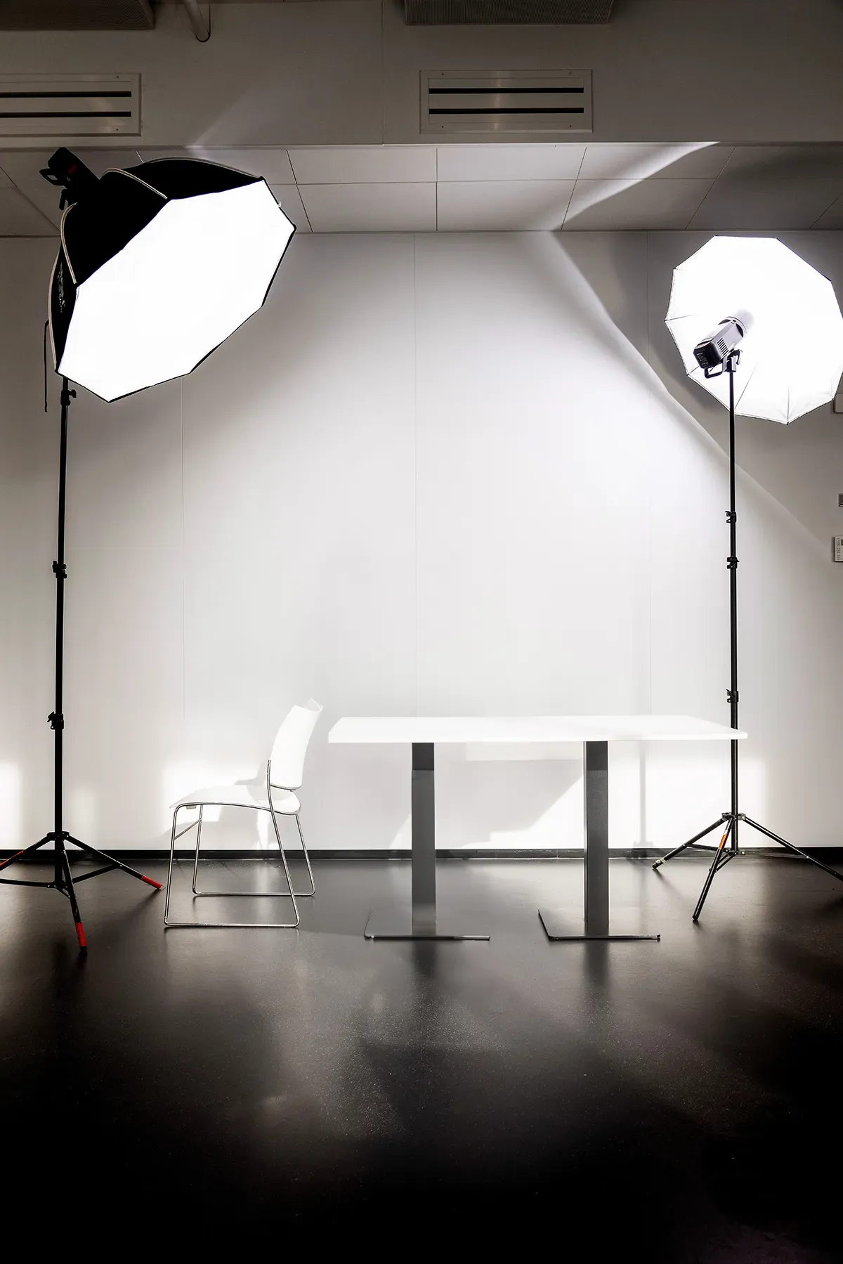 Een minimalistische fotostudio-opstelling met een witte stoel, een rechthoekige tafel en twee grote softboxlampen op standaards aan weerszijden, die helder licht op de scène werpen. De werkwijze van de studio benadrukt effen witte muren en een donkere, reflecterende vloer voor optimale verlichting en helderheid.