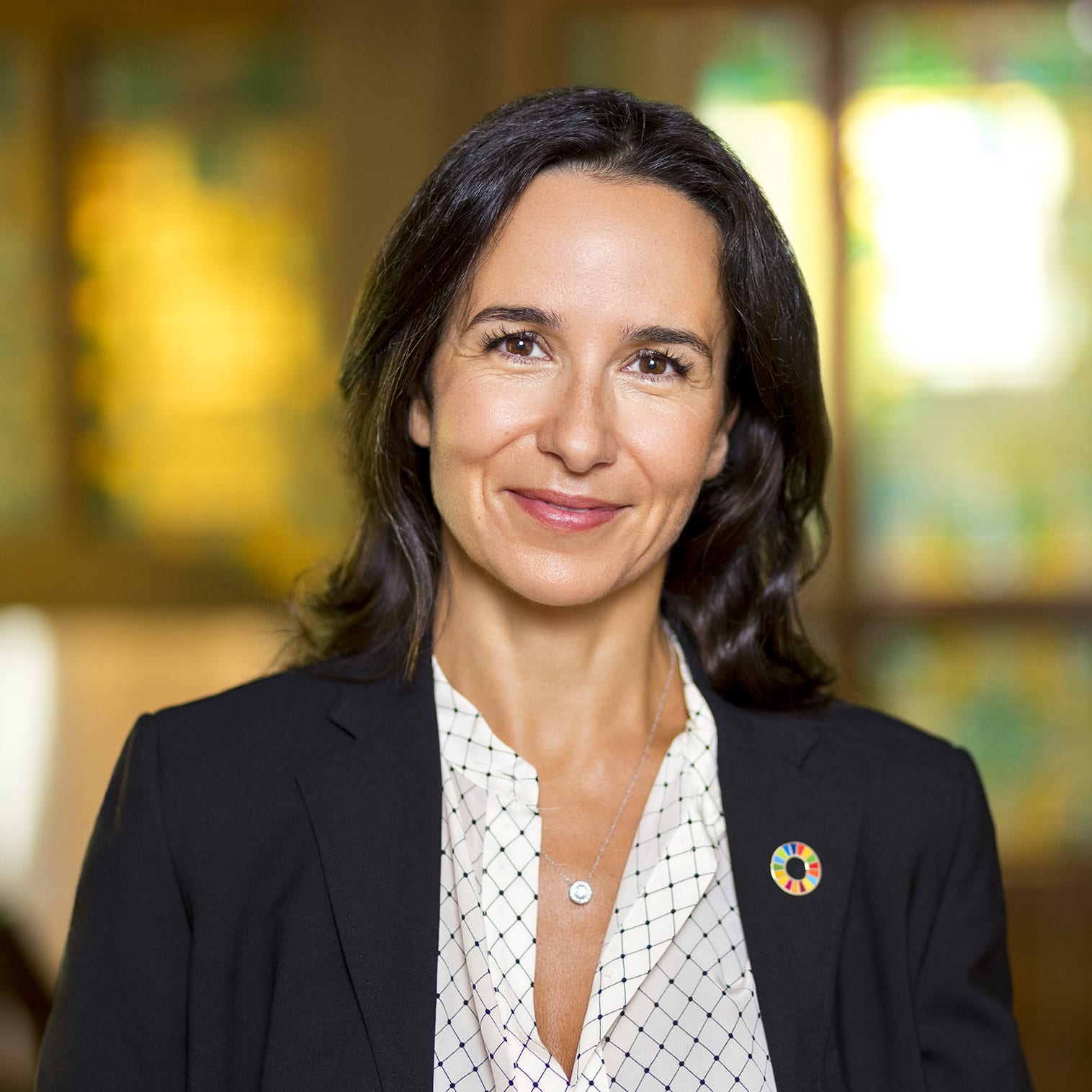 Een vrouw met lang, donker haar lacht naar de camera. Ze draagt een donkere blazer en een witte blouse met een rasterpatroon. Ze heeft een speld met kleurrijke cirkelsegmenten op haar revers. De achtergrond, zacht vervaagd door wat lijkt op glas-in-loodpanelen, voegt een professionele toets toe die je vaak ziet in zakelijke portretten voor haar portfolio.